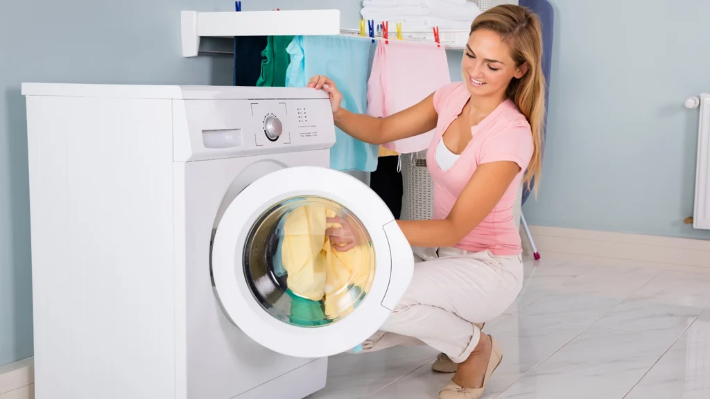 Smiling woman kneeling on the floor, loading colorful laundry into a front-loading washing machine in a bright, clean laundry room with clothes drying on a rack in the background.